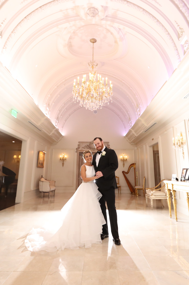 Bride in a tiered organza gown with a belt and chapel train, poses with the groom under a crystal chandelier in a ballroom.
