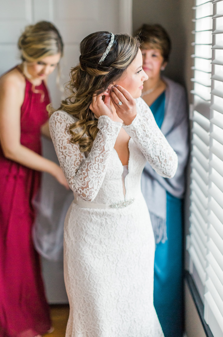 Bride in a long sleeved lace gown with a plunging neck and beaded belt detail,  gets ready with her bridesmaid by a window.