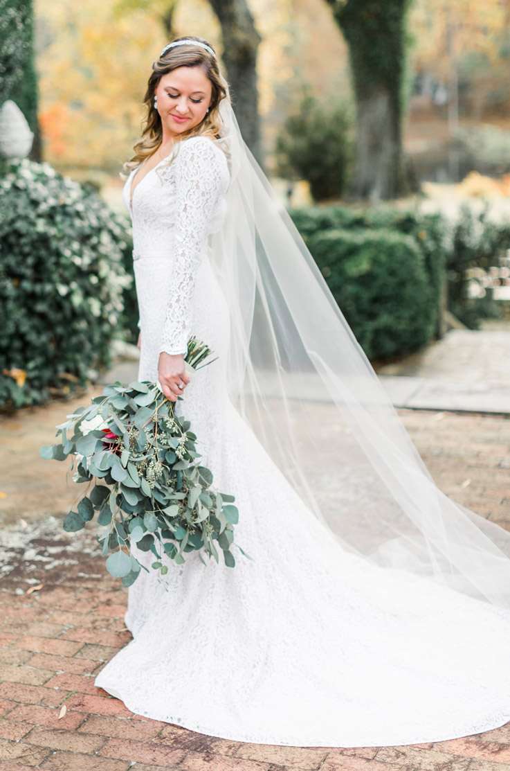 Bride in a lace fit and flare wedding dress with a chapel train and veil, posing and holds a bouquet in a garden setting.
