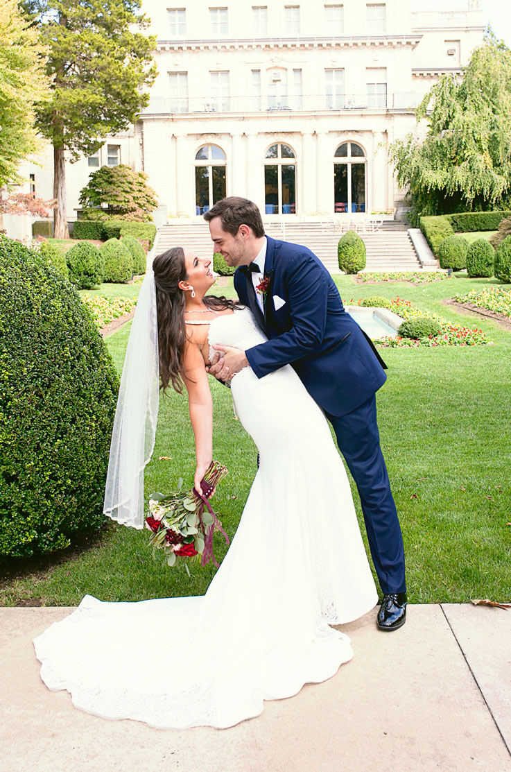 Groom dips the bride in a crepe fit and flare gown with a lace bodice and veil, holding a bouquet in front of the venue.