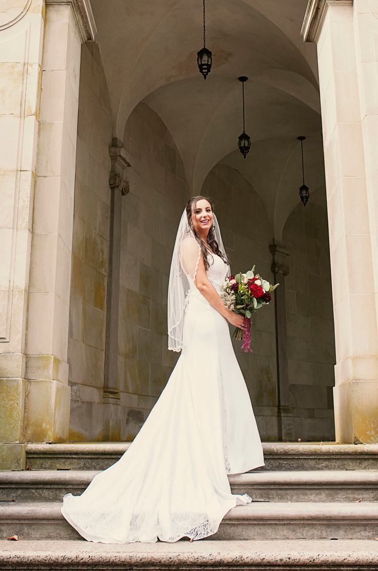 Bride stands on a grand stone staircase in an archway, wearing a sweetheart fit and flare gown with a veil and chapel train. 