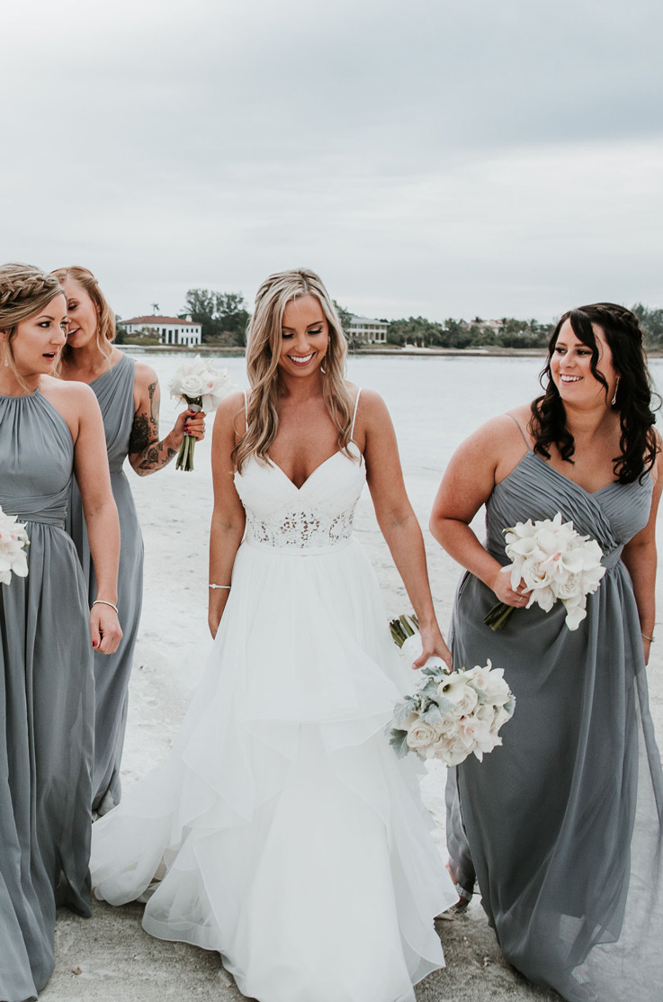 Bride in a lace bodice gown with a V-neck and a layered tulle skirt, walks down a beach with her bridesmaids holding bouquets.