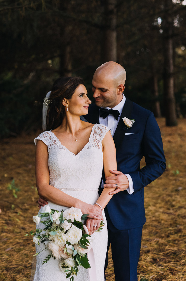  Bride in a lace fit and flare gown with cap sleeves and a veil, stands with the groom in an embrace outdoors holding a bouquet.