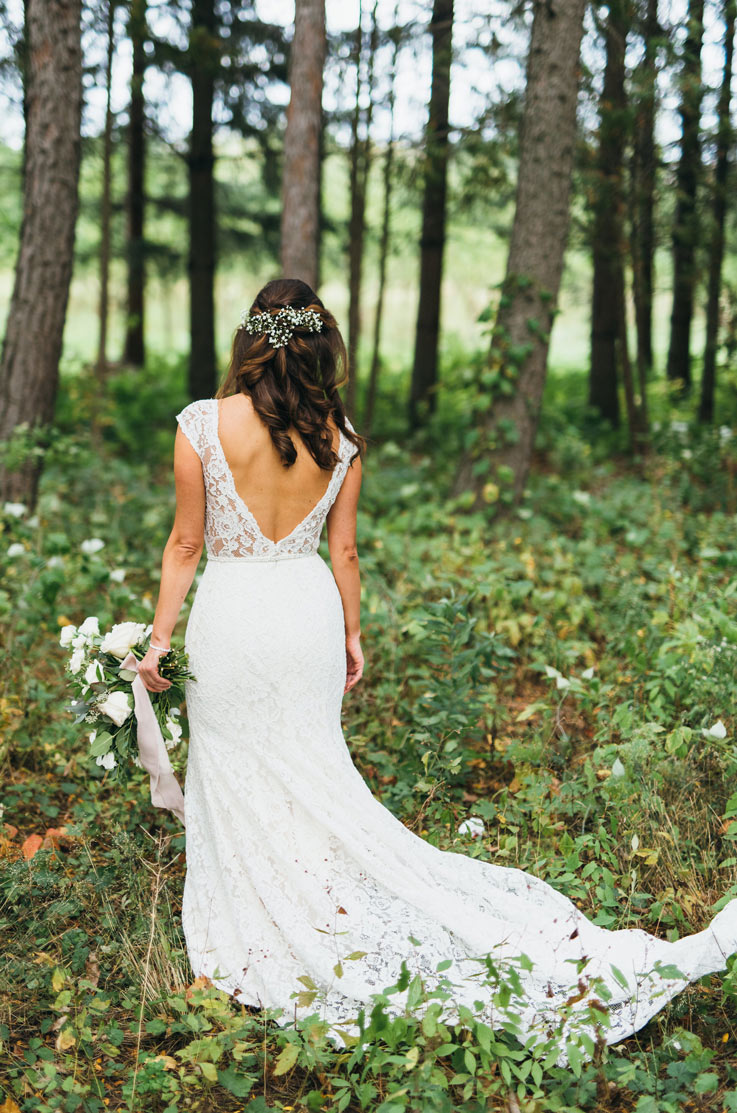 Back of bride in a low open back lace gown with a fit and flare skirt, stands in a forest and holds a white bouquet