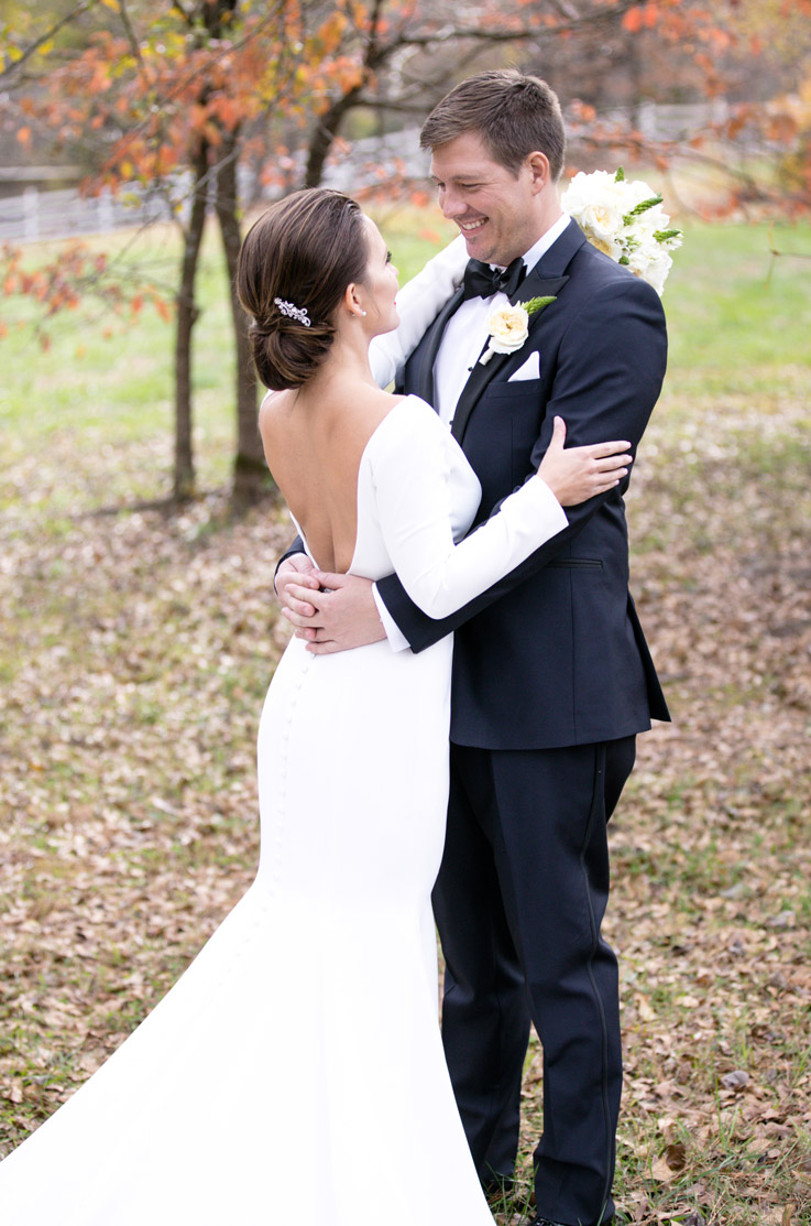 Bride in a fit and flare crepe gown with a low back and long sleeves, faces groom in an embrace in a outdoor fall setting.