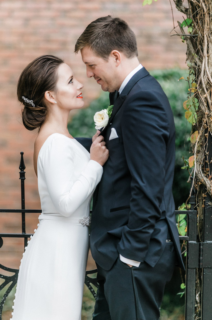 Bride in a boat neck long sleeve wedding dress with an open back and belt detail, faces groom in an embrace by an iron fence.