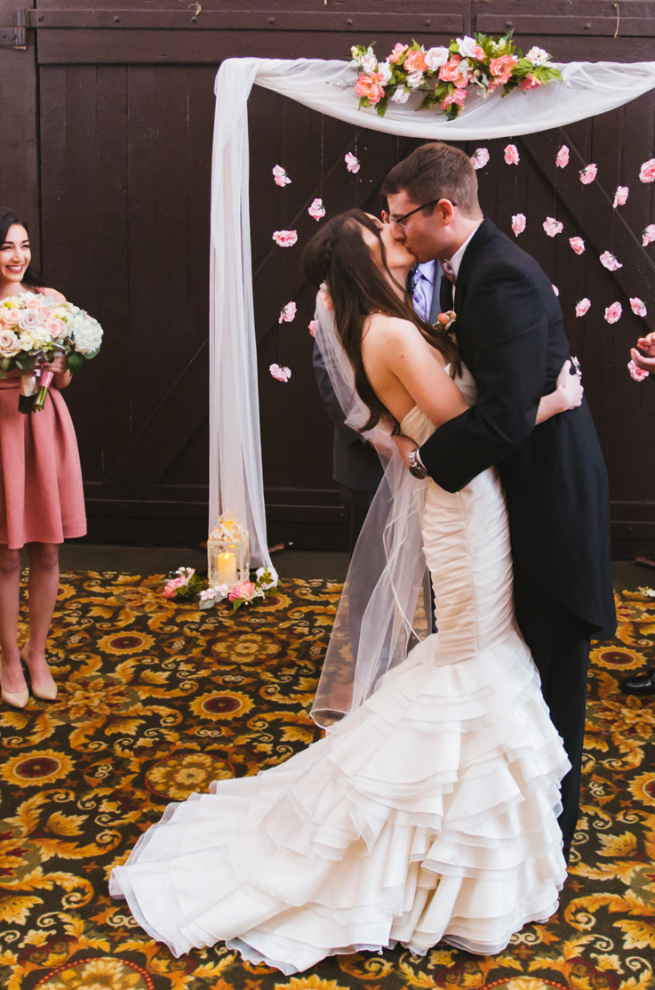 Bride in a ruffled taffeta and organza gown with a tiered skirt and veil, kisses groom under a floral arch at the altar.