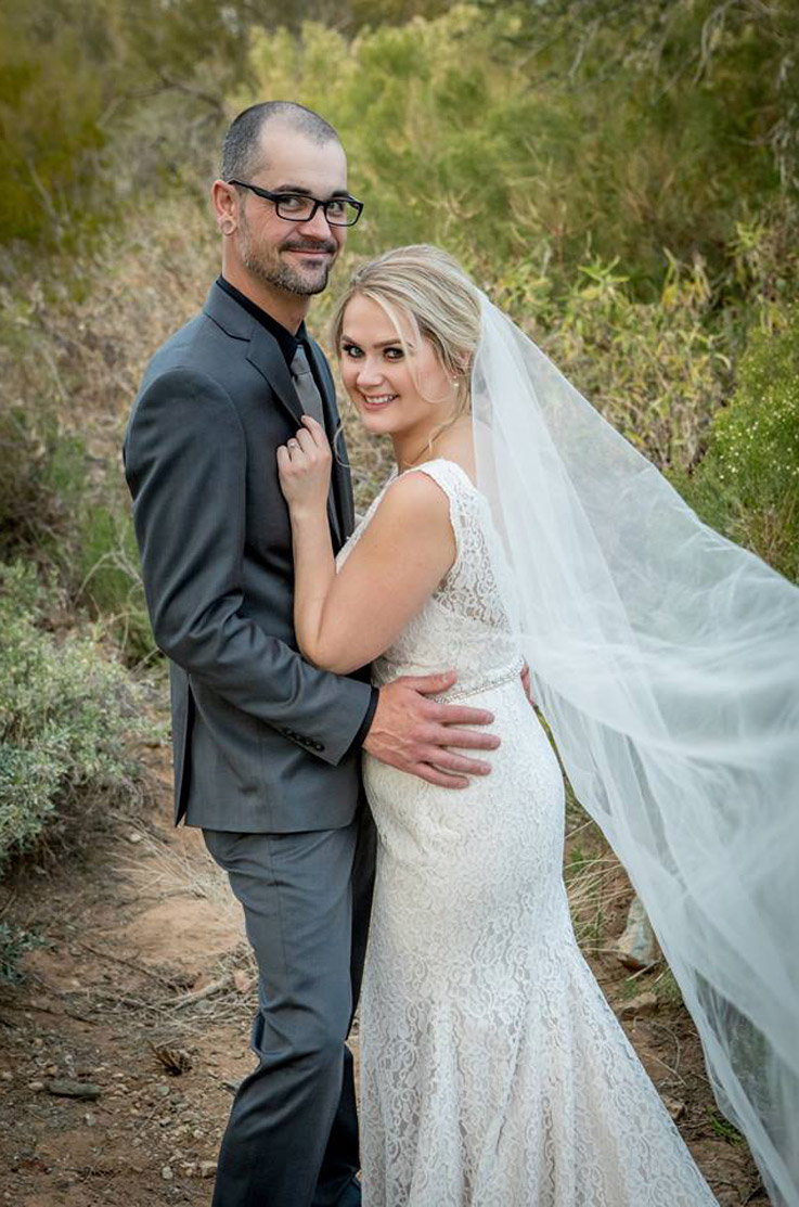 Groom embraces bride in a sleeveless fit and flare gown in lace with a flowing veil, and they stand in a desert.