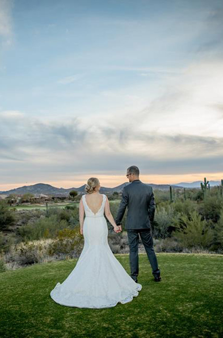 Back view of groom holding hands with bride in a lace fit and flare gown with a scalloped neckline, looking out at a desert.