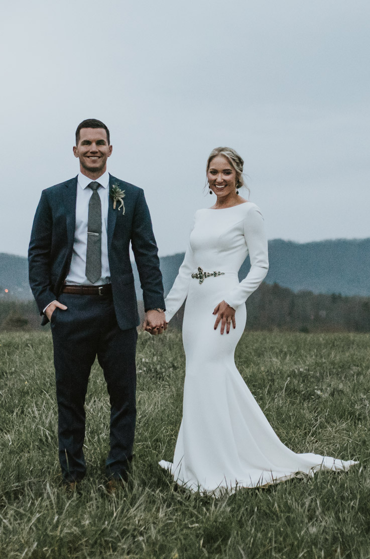 Bride in a bateau neck crepe wedding gown with long sleeves and a belt, smiles and holds hands with groom on a mountaintop.