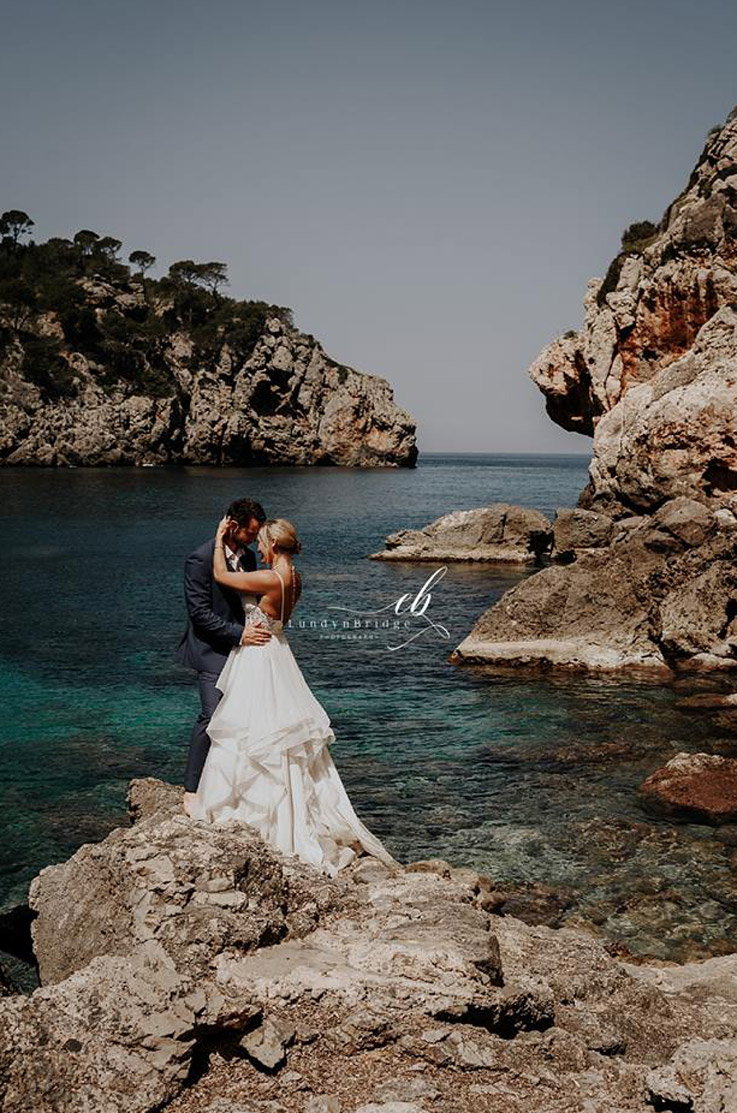 Bride in a layered tulle wedding dress with lace, embraces groom on a rocky seaside cliff, in front of turquoise water.