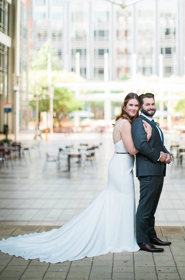  Bride in a crepe fit and flare gown with crossover straps and a belt, embraces groom in a city courtyard.