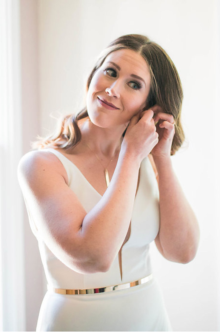 Close-up of bride smiling in a sleeveless crepe wedding gown with a plunging neck and gold belt, getting ready indoors.