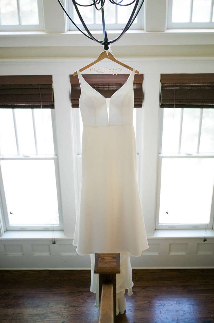 A sleeveless fit and flare wedding dress in crepe with a plunging V-neckline, hangs in a bright bridal suite by windows.