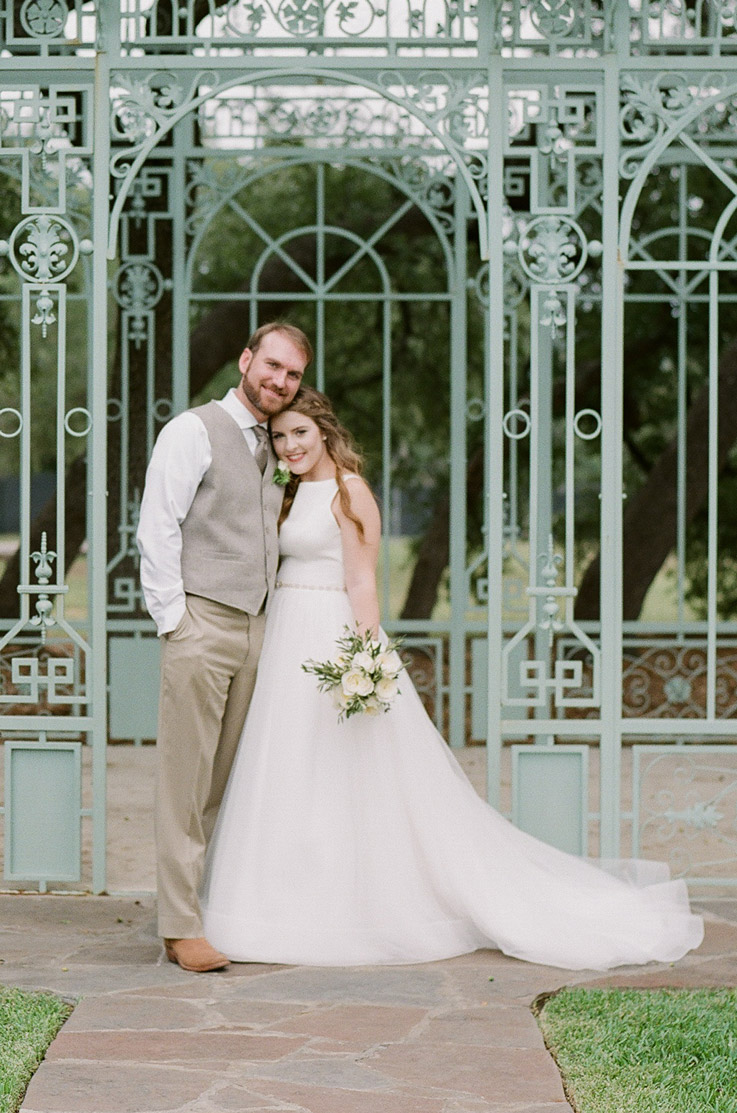 Bride in a high neck crepe gown with a tulle skirt, holding a bouquet and stands next to the groom by a wrought iron gazebo.