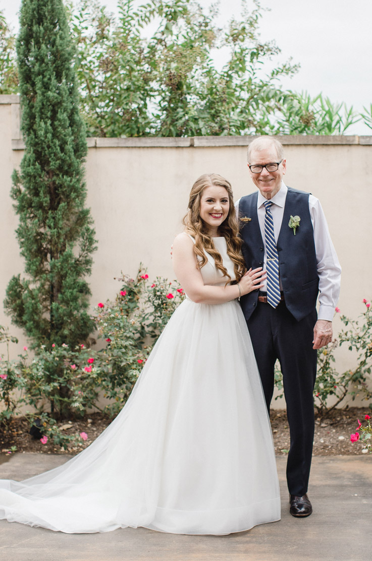 Bride stands next to the groom in a sleeveless tulle gown with a crepe bodice by a wall with rose bushes.