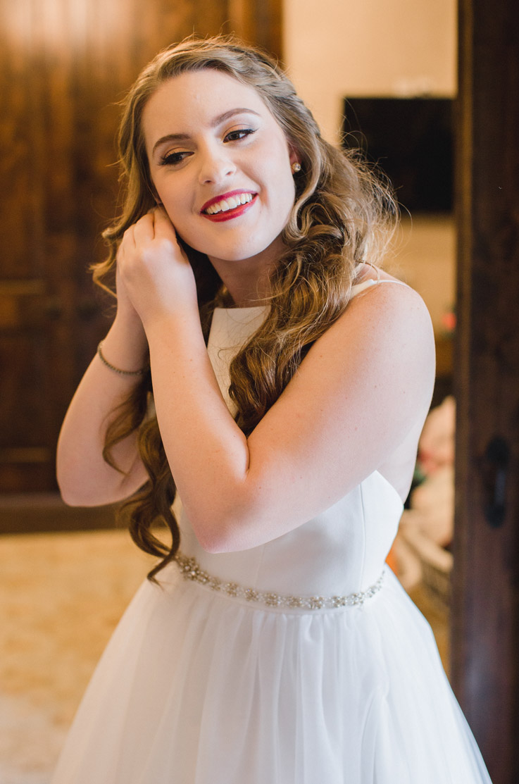 Bride in a sleeveless crepe wedding gown with a tulle skirt and beaded belt, smiles while getting ready indoors. 