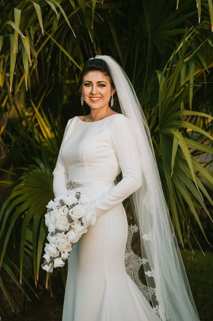 Smiling bride in a fit and flare gown with a long veil, sleeves and belt detail, holding a white bouquet by palm trees.