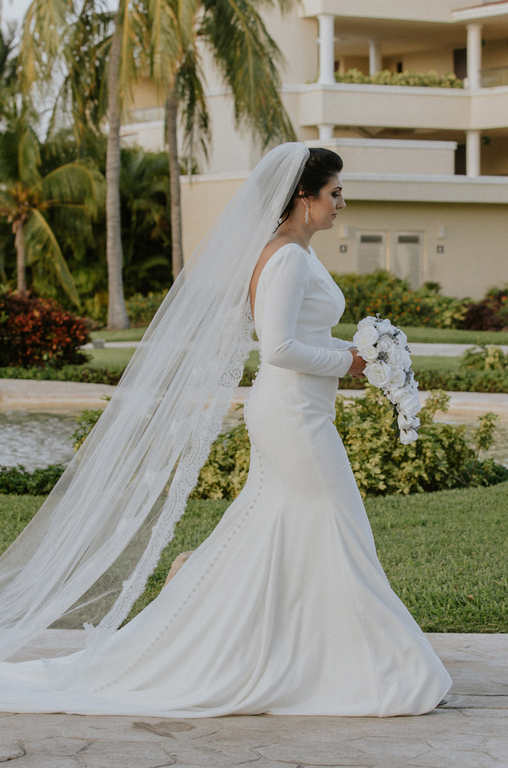 Bride walks through a tropical resort in a fit and flare crepe wedding gown with a bateau neckline, sleeves and a veil.