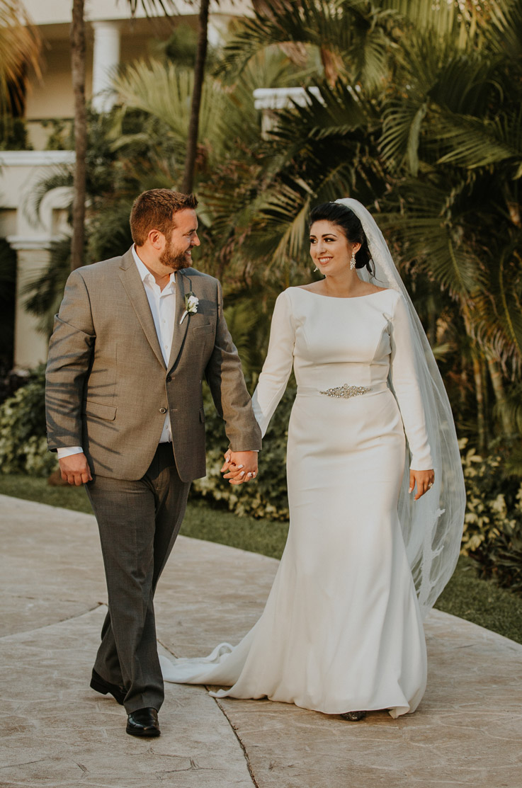 Bride walks hand in hand with groom in a bateau neck wedding dress in crepe with a veil and long sleeves, and they are smiling.