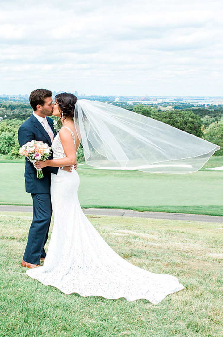 Bride in a lace fit and flare gown with a veil, stands on a grassy hill holding a bouquet and kisses groom in a blue suit.