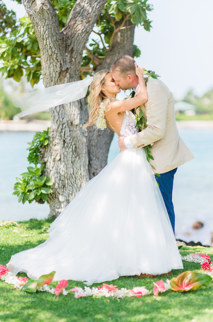Bride in a flowing tulle wedding dress with a lace bodice and lei, kisses the groom by a shoreline under a tree.