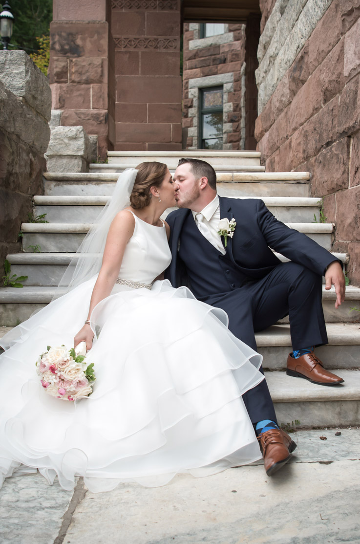 Groom kisses the bride in a bateau neck tiered gown with a satin bodice, belt and a veil, holding a bouquet on stone steps.