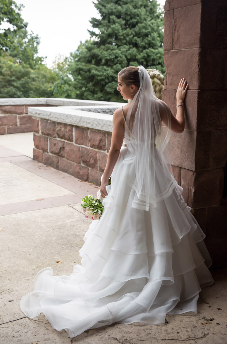 Bride in a tiered organza wedding gown with a chapel train and veil, poses by a stone building and holds a bouquet.
