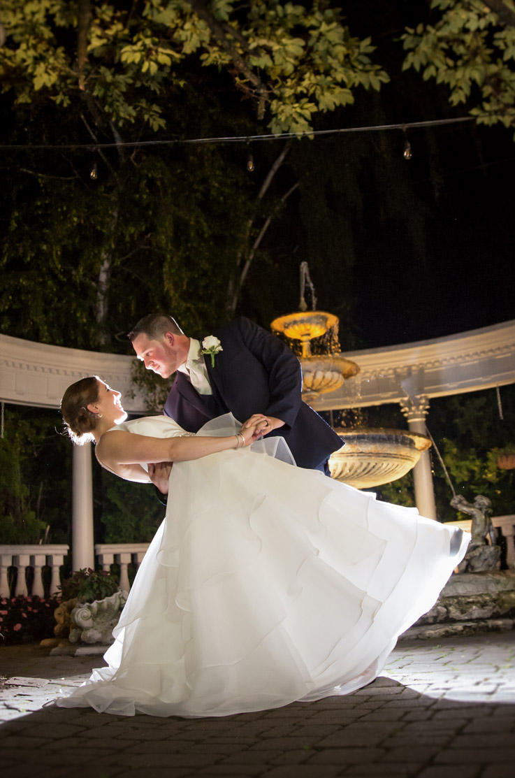 Groom dips his bride in a tiered organza ballgown with a chapel train and satin bodice, at ornate fountain at night.