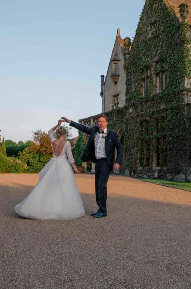 Groom twirls his bride in a tulle gown with a low open back and lace bodice with sleeves, outside an ivy-covered manor.