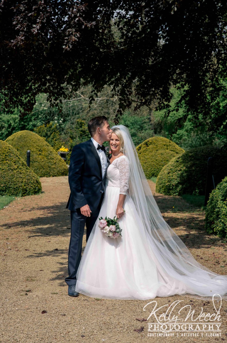 Bride in a lace bodice gown with a full tulle skirt, a chapel train and veil, smiling and stands next to groom with flowers. 