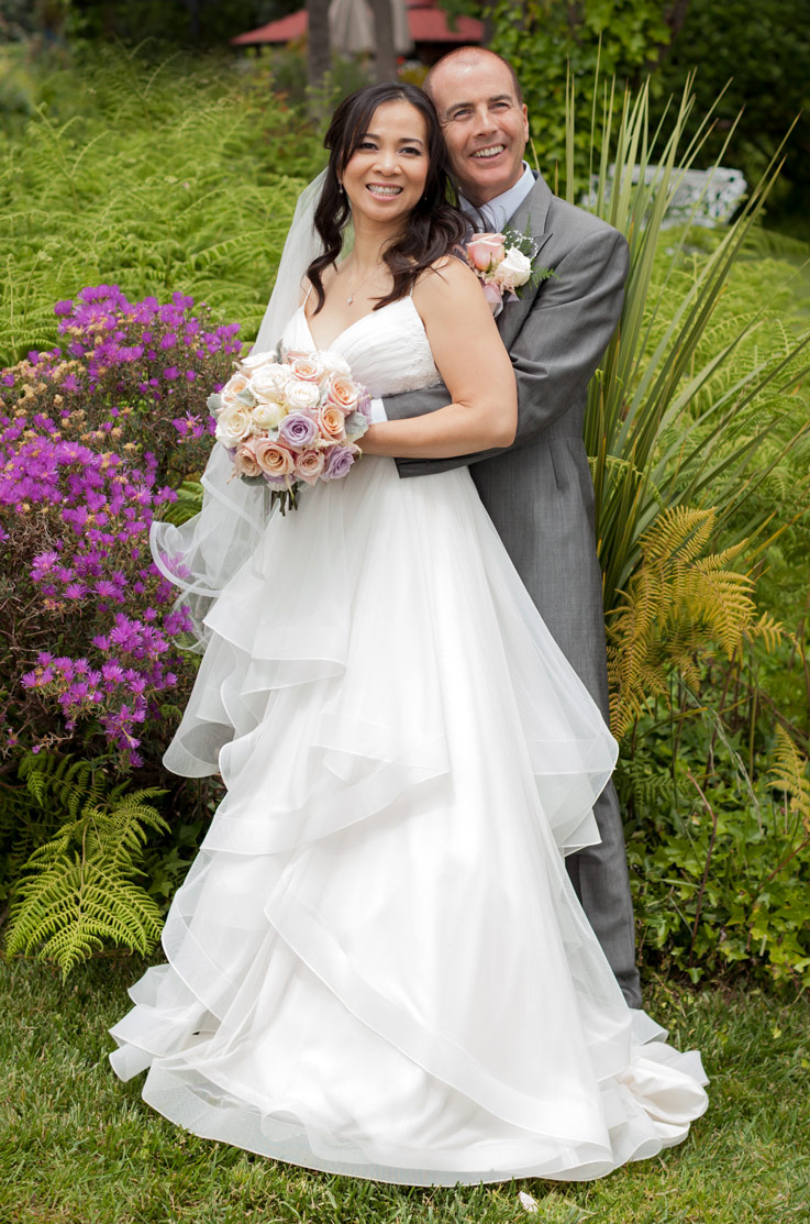 Groom embraces bride in a tulle layered wedding gown with a V-neck and veil, holding a bouquet in a garden setting.