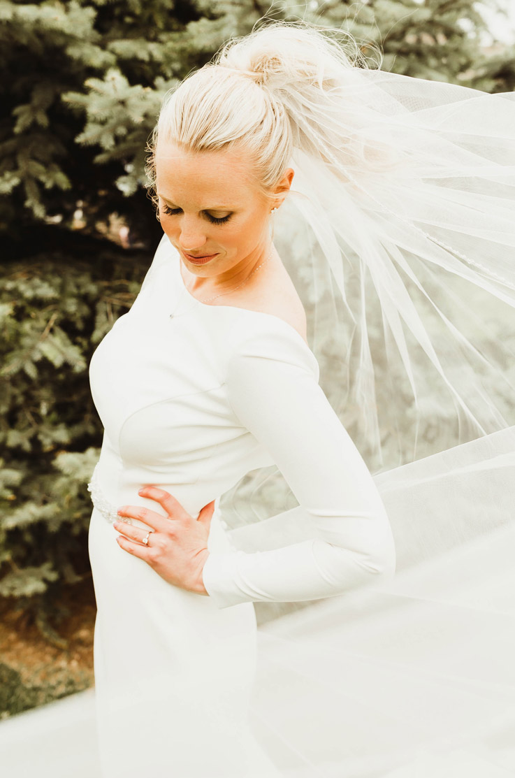 Close-up of bride in a bateau neck crepe wedding dress with long sleeves and a veil, poses outdoors among evergreen trees.