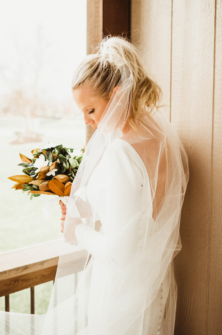 Back of bride in a long-sleeve crepe gown with a bateau neck, a veil and a buttoned back, holding a bouquet indoors.