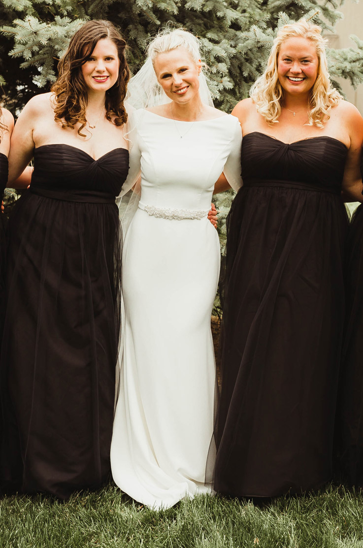 Bride in a long sleeve crepe gown with a belt, bateau neck and veil, smiles next to bridesmaids in black dresses by pine trees.