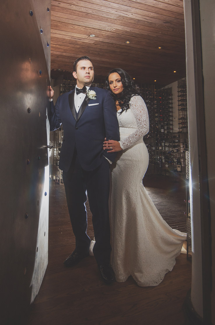 Bride in a lace long sleeve gown with a fit and flare skirt, poses with the groom in a tuxedo in a doorway to a cellar. 