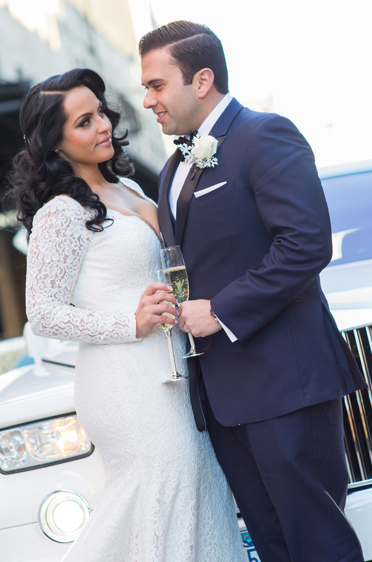Bride in a lace fit and flare wedding dress with sleeves and a plunging neck, faces groom and they hold champagne glasses.
