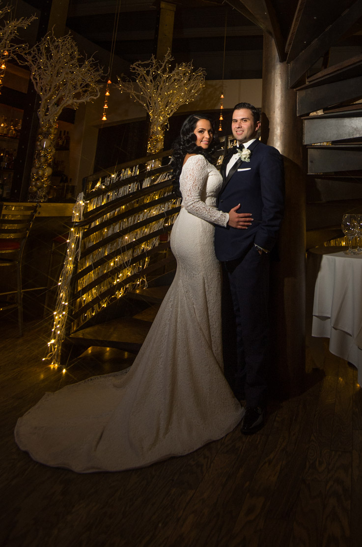 Bride stands beside groom in a lace fit and flare gown with sleeves at a winding spiral staircase with dramatic lighting.