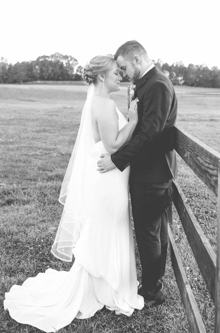 Groom embraces bride in a crepe wedding gown with thin straps and a low back, as they face each other by a fence in a field.