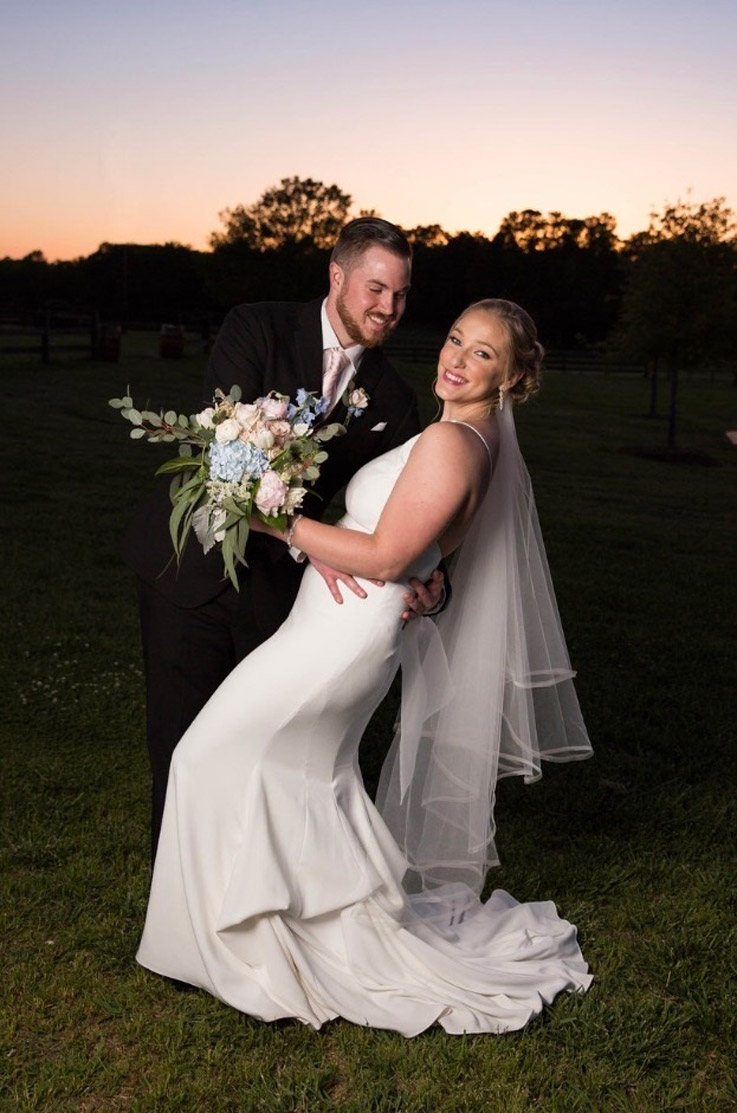 Groom dips his bride in a crepe fit and flare gown with a veil, in the countryside at sunset and she holds a bouquet.