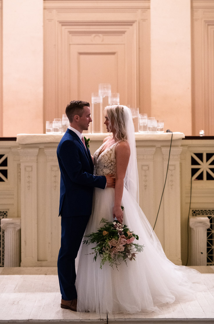Bride in a V-neck lace gown with a flowing tulle skirt and veil, holds a large bouquet and faces her groom near candlelight.