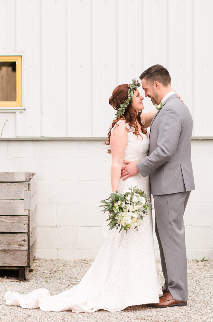 Bride in a low open back lace gown with a scalloped neckline and sash with a tie, embraces groom in a gray suit.