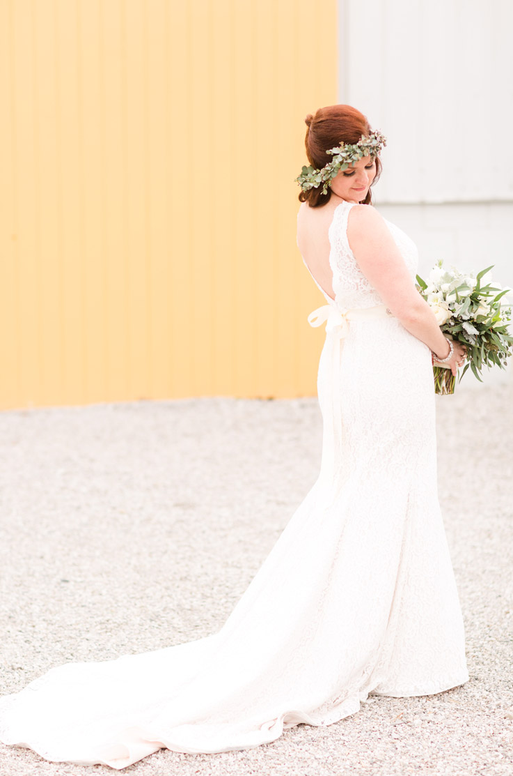 Bride in a low open back lace gown with a sash, flower crown and chapel train, poses outside with a bouquet by a bright wall. 