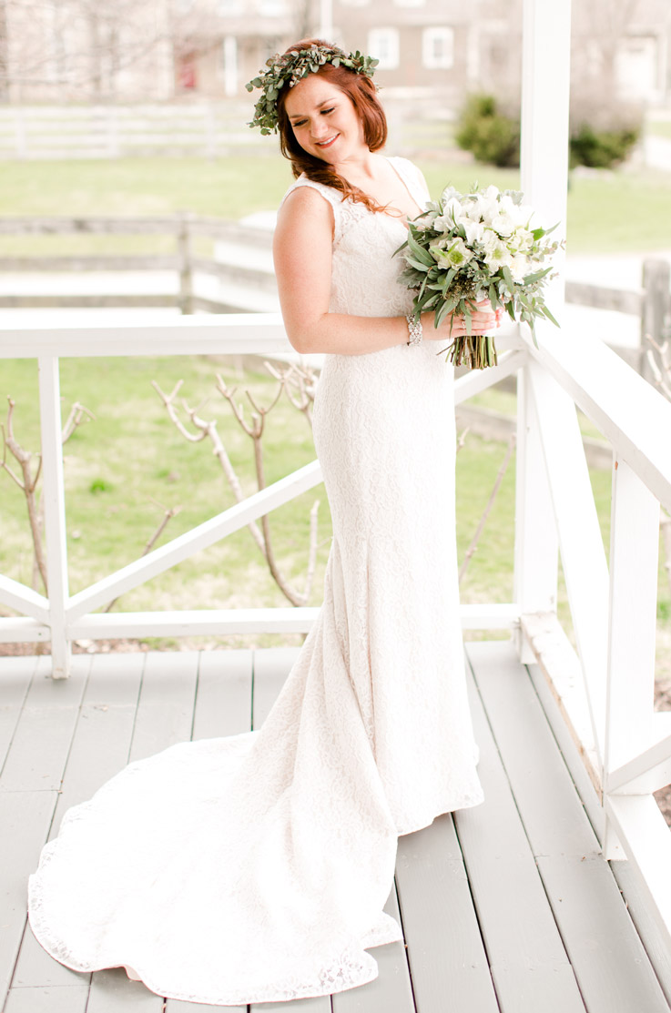 Bride in a lace fit and flare wedding dress with a chapel train and plunging neck, holds a bouquet on a white farmhouse porch.
