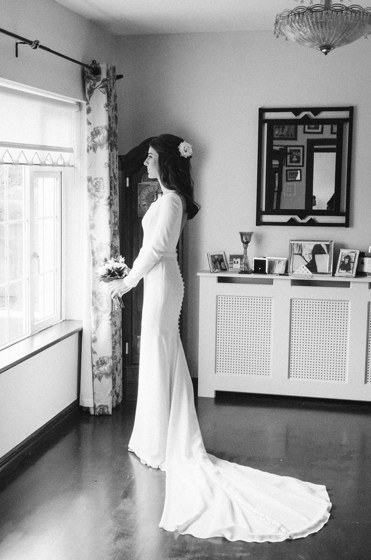 Bride stands by a window in a fit and flare crepe gown with long sleeves and a buttoned back, indoors in black and white.