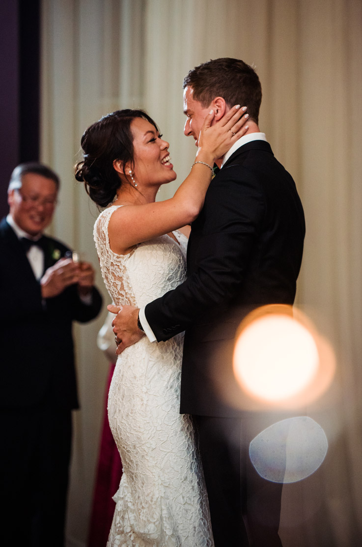 Bride in a fit and flare lace gown with cap sleeves, embraces groom and they smile at each other during reception.