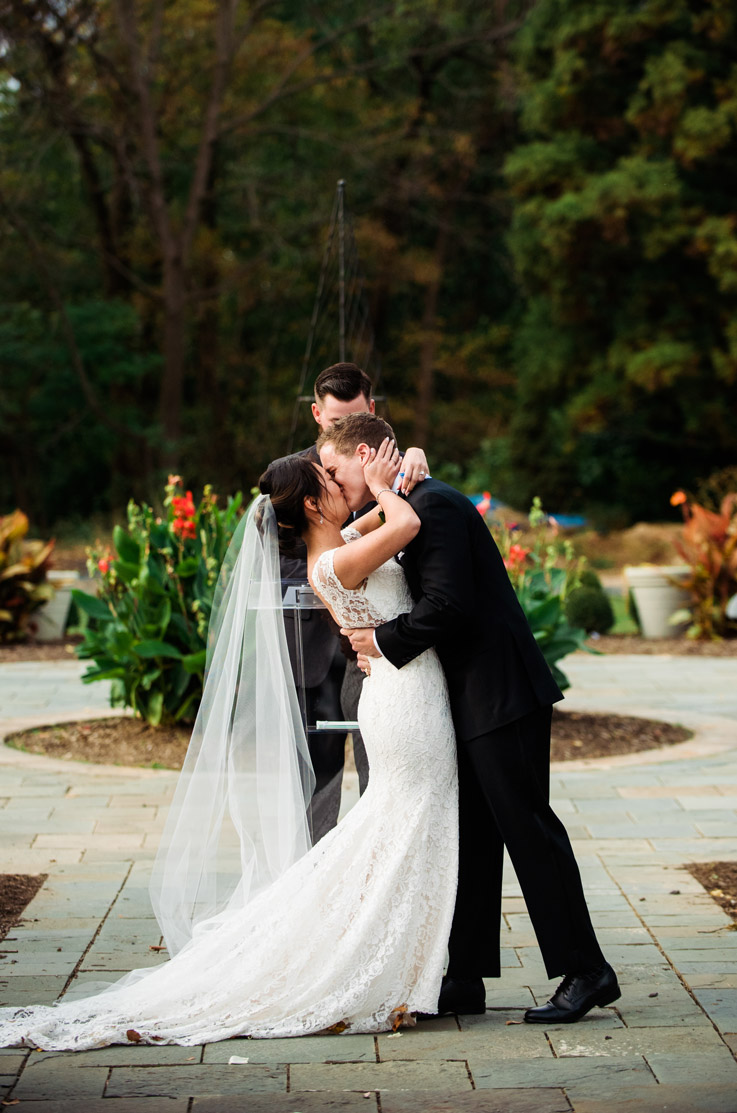 Bride in a lace fit and flare wedding gown with cap sleeves and a veil kisses groom in a black tux at outdoor garden ceremony. 
