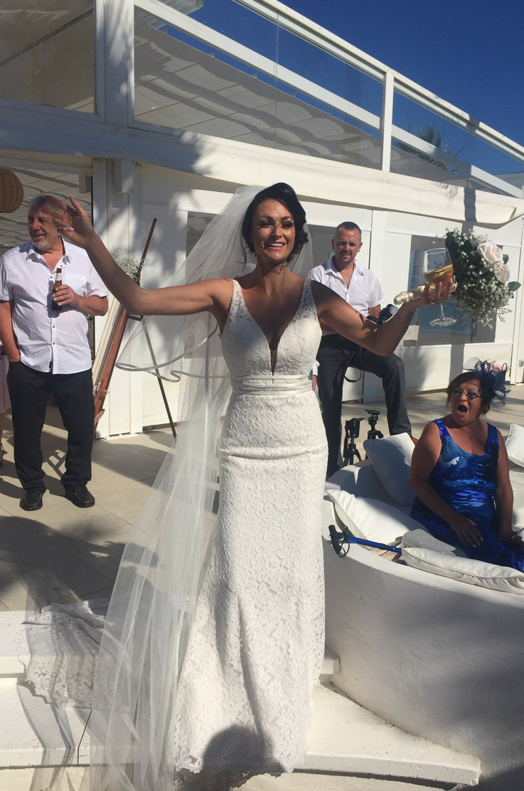 Bride in a lace fit and flare wedding dress with a plunging neck and veil,  is celebrating at a sunny seaside venue.