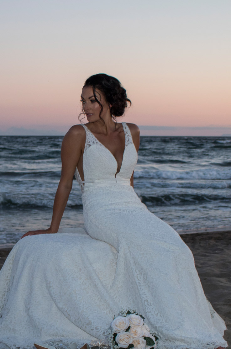 Bride sits on a beach by the shore in a lace fit and flare gown and looks to the side, with a bouquet of roses at sunset.