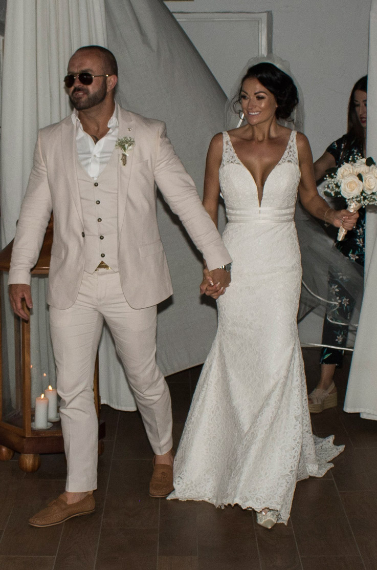 Bride holds hands with groom and is wearing a lace fit and flare gown with a deep-V neck indoors and is holding a bouquet. 